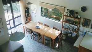 Een eetgedeelte met een houten tafel en stoelen, een raam met uitzicht op groen, en een keuken met planken en een klok.