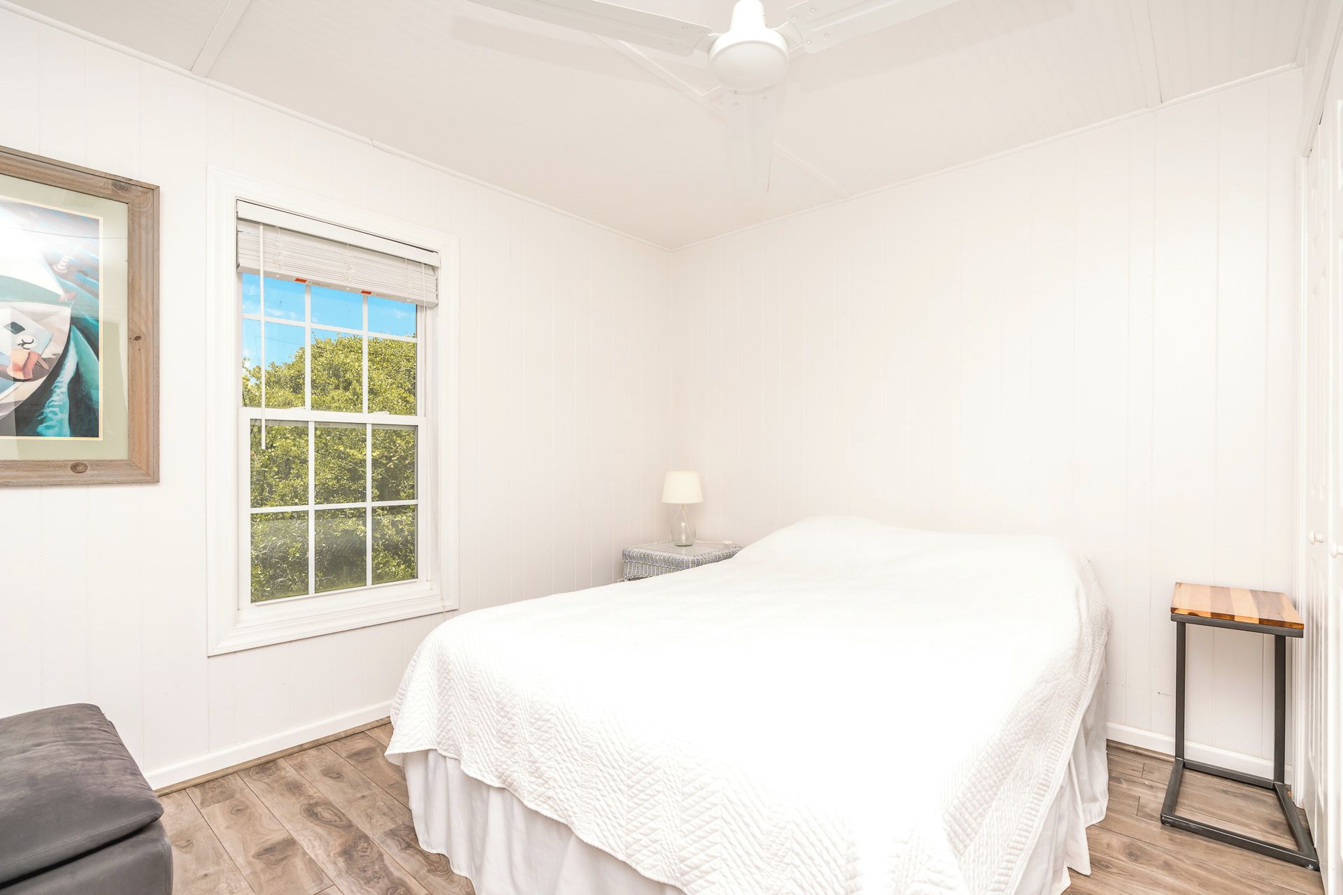 A bedroom with a bed, a window, a bedside table with a lamp, and a small wooden stool.