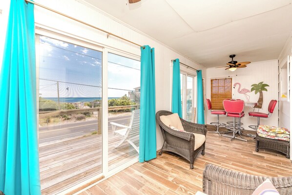 A balcony with a wicker chair, a dining table with red chairs, and a view of the road and greenery. - Ruff Life - Classic Island Cottage - 4 BR and Easy Beach Access. Dog Friendly! (Surf City)