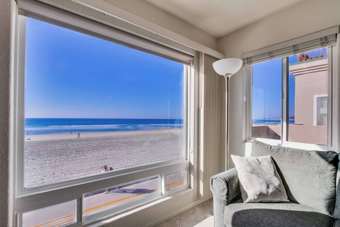 A room with a large window overlooking a beach, a sofa with cushions, and a floor lamp.