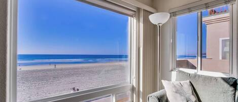 Ein Zimmer mit großem Fenster, das auf einen Strand blickt, einer Couch mit Kissen und einer Stehlampe.