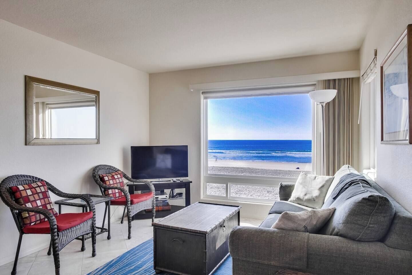 A hotel room with a television, two wicker chairs, a sofa, and a large window offering a beach view.