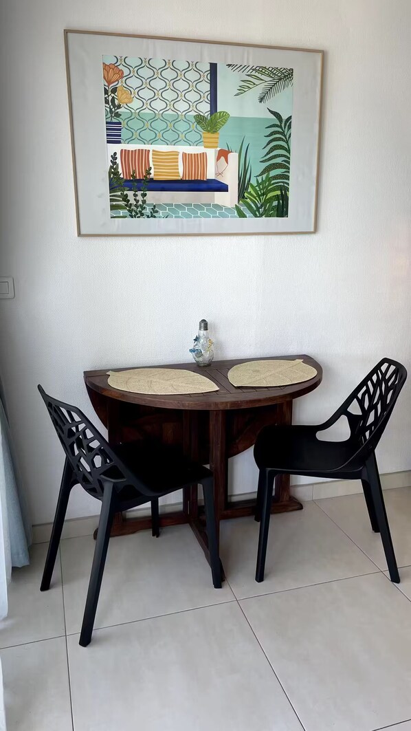 A dining area with a round wooden table, two black chairs, and a framed artwork above. - Superb Beachfront Studio with Ocean View in Marigot (Sandy Ground)
