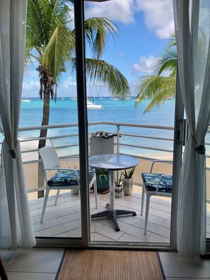 A balcony with a table and chairs, overlooking a beach with palm trees and boats. - Superb Beachfront Studio with Ocean View in Marigot (Sandy Ground)