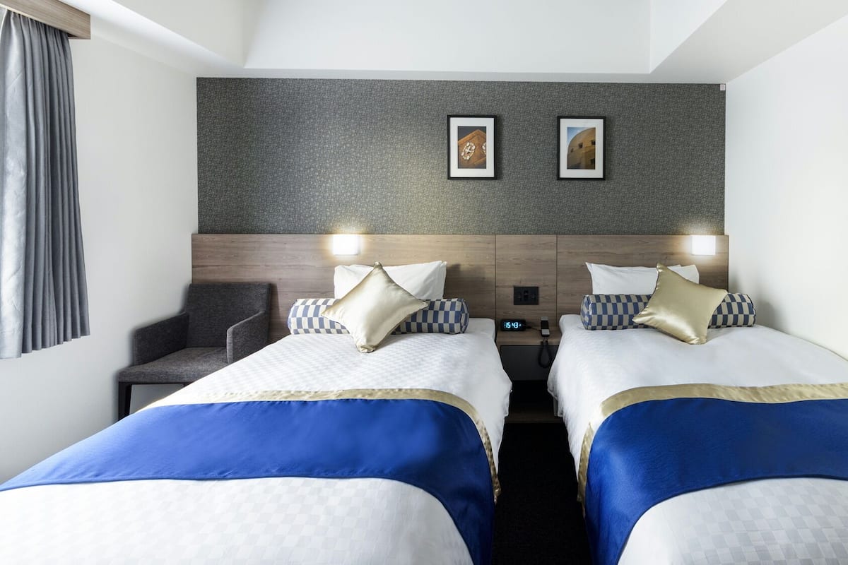 Habitación de hotel con dos camas, una silla, un reloj y dos cuadros en la pared.