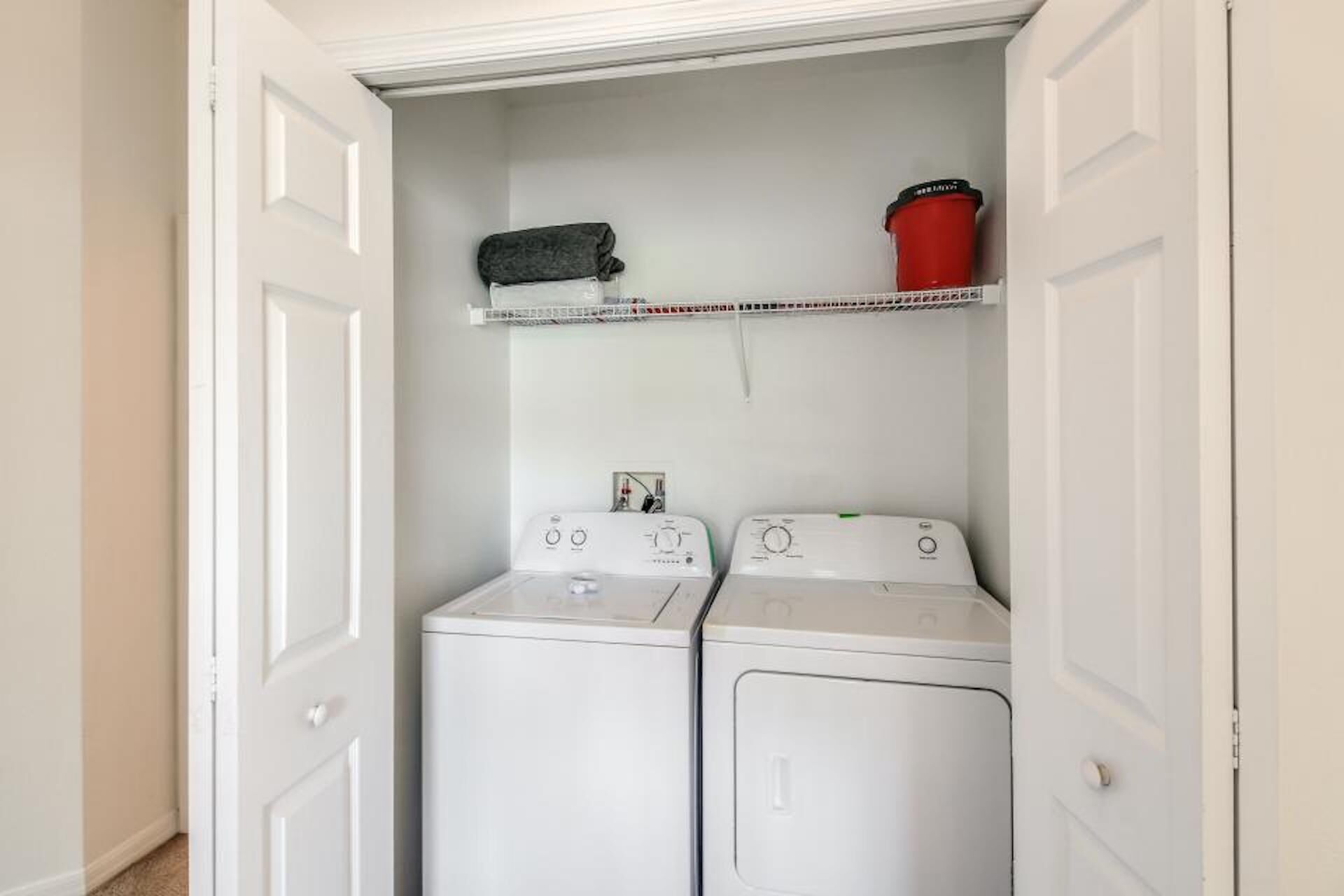A laundry room with two white front-loading washers and dryers, a shelf above with a black bag and a red bucket, and white doors.