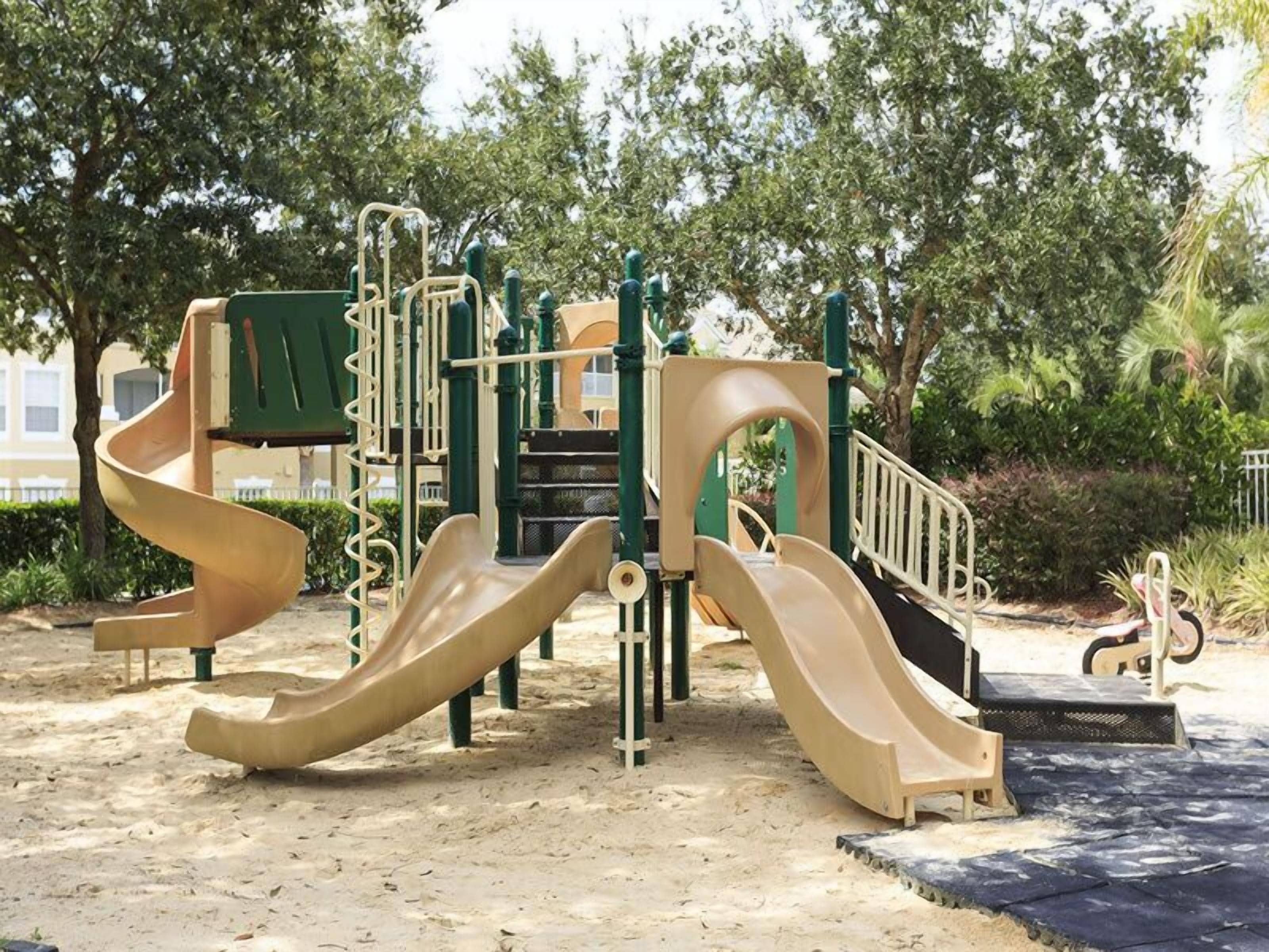 Ein Spielplatz mit mehreren Rutschen und Klettergerüsten, umgeben von Bäumen und einem Sandboden.