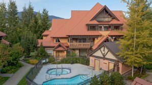 A large building with a swimming pool surrounded by a fence, set in a wooded area with mountains in the background.
