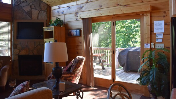 A cozy cabin interior with a stone fireplace, a comfortable seating area, and a large sliding glass door leading to an outdoor deck. - MTN CABIN LAKEFRONT,CANOE/3 KAYAKS,POOL TABLE,DOCK,FIRE PIT, FIREPLACE,JACUZZI (Butler)