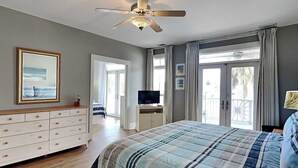 A bedroom with a bed, a dresser, a ceiling fan, a television, and a balcony with curtains.