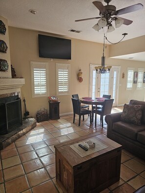 A living room with a fireplace, a ceiling fan, a TV mounted on the wall, and a dining area with a table and chairs.