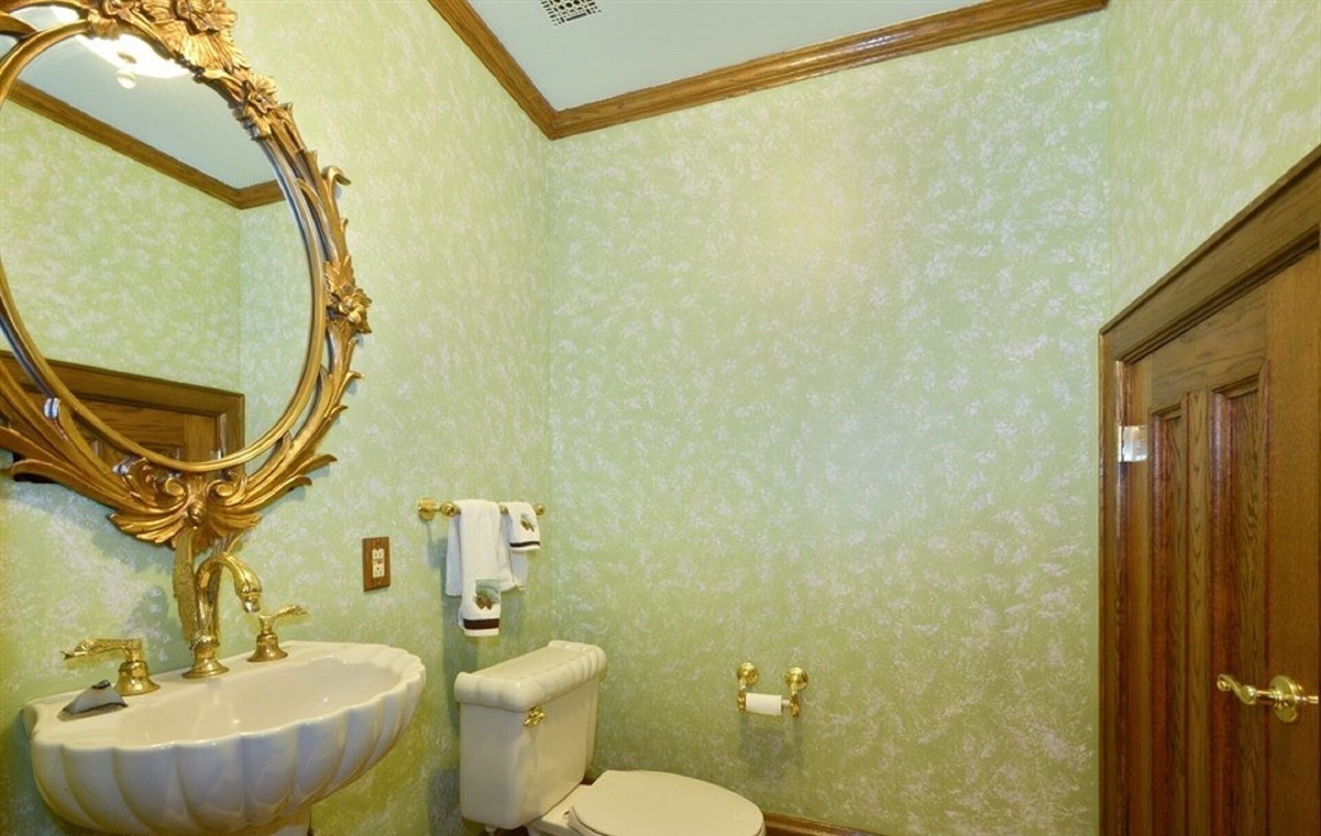 A bathroom with a large ornate mirror, a white sink with gold fixtures, a toilet, and a wooden door.