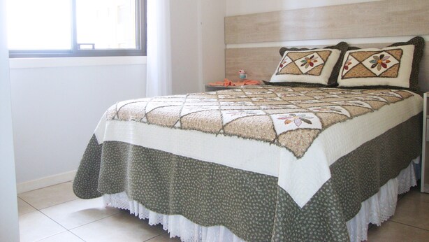 A neatly made bed with a quilt, a bedside table with a lamp, and a window with curtains. - Inactive ad (capao da canoa)