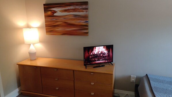 A wooden dresser with drawers, a television on top displaying a fire scene, a wall-mounted painting, and a lamp on the dresser.