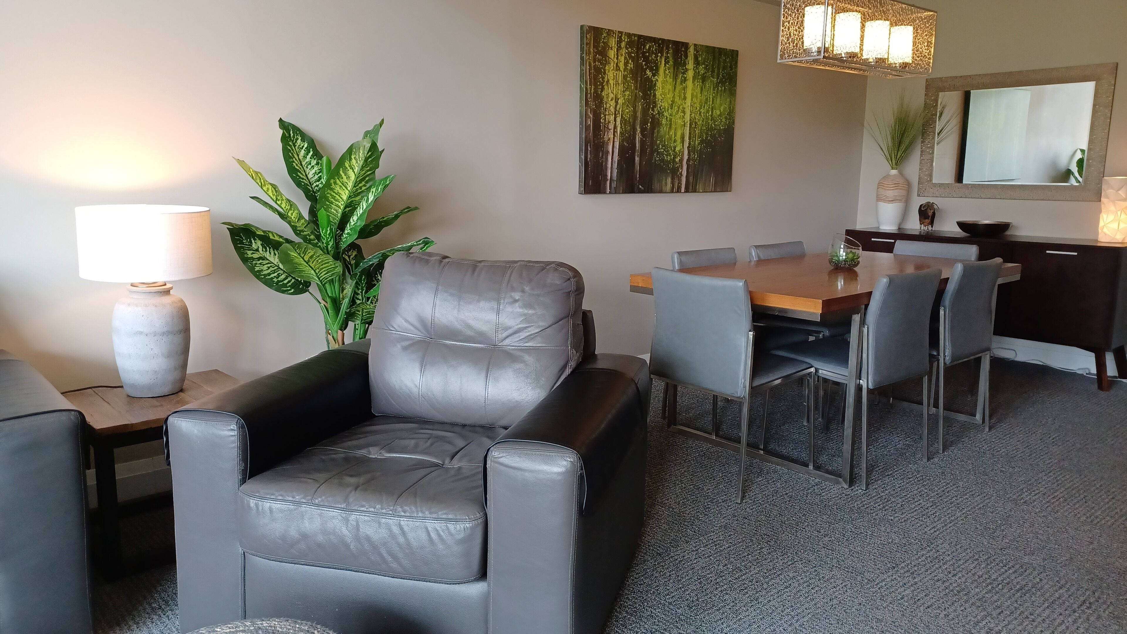 Ein modernes Wohnzimmer mit Ledersofa, einem Esstisch mit Stühlen, einer großen Kübelpflanze und einem Bild an der Wand.
