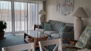 Una sala de estar con un sofá, una mesa de centro y una mesa de comedor con sillas. Hay dos lámparas sobre el sofá y una planta en maceta cerca de la ventana.