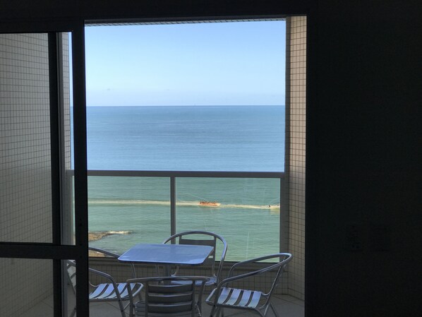Un balcón con mesa y sillas, con vista a una playa donde se ve un barco en el agua.