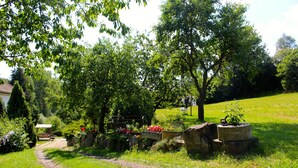 Un sentier de jardin mène à une structure en bois dotée d’un banc, le tout étant entouré d’une végétation luxuriante et de plantes en fleurs.