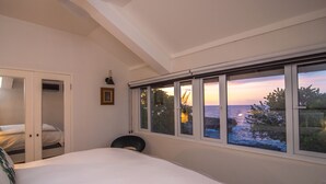 Un dormitorio con una cama grande, una silla, un armario y una ventana con vista al mar y al atardecer.