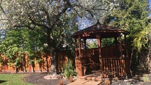 Un pabellón con techo de madera y lados abiertos, rodeado por un jardín con diversas plantas y un árbol grande con flores blancas.
