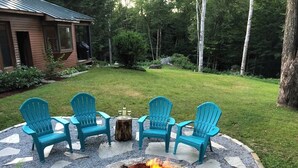 A fire pit with four blue Adirondack chairs arranged around it, set in a stone circle on a grassy lawn with a wooden deck and a house in the background. - Snowberry Hill, 5 bedroom, Private Luxury Ski Home on Killington Mtn, sleeps 12 (Killington)