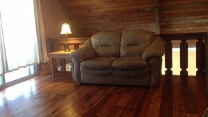 A wooden-floored room with a leather sofa, a small table with a lamp, and a window with curtains. - Lakeside barn wood cottage just remodeled with all the modern Conveniences! (Greentown, PA)