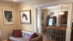 Uma sala de estar com sofá, uma mesa de jantar com cadeiras e uma cozinha ao fundo.
