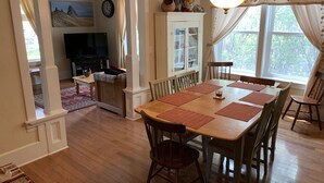 Uma sala de jantar com mesa e cadeiras de madeira, televisão ao fundo e vista para o exterior através de grandes janelas.