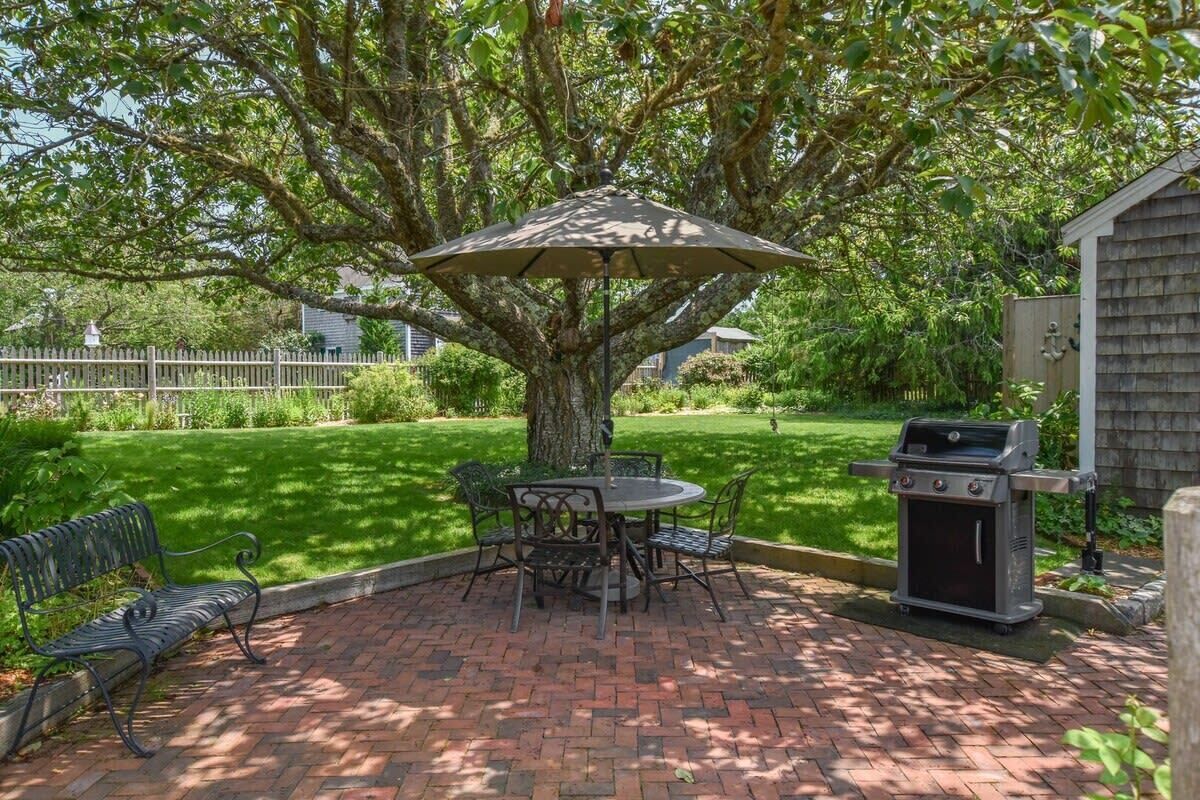 A patio area with a brick floor, a round table with chairs, a grill, and a tree providing shade.