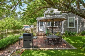 A patio with a grill, outdoor table, and chairs, surrounded by a wooden fence and lush greenery.