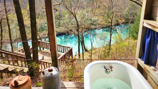 A wooden deck overlooking a turquoise lake with a rustic bathtub and a metal barrel. - Secluded River Cabin Retreat with Outdoor Bathhouse and Kayaks on 80 acres! (Caddo Gap)