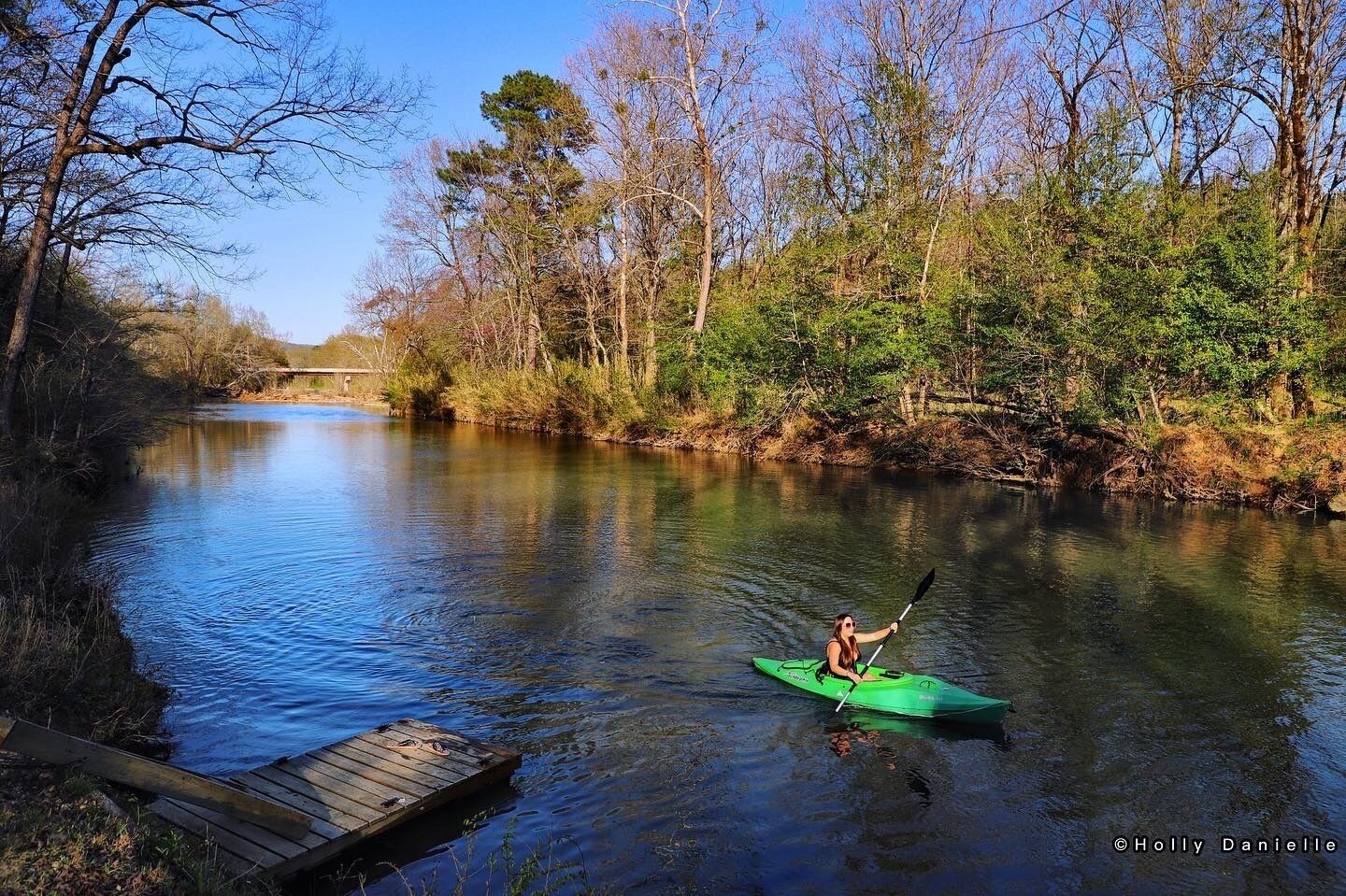 A kayaker on a river with a wooden dock and trees in the background.