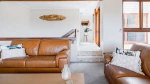 Uma sala de estar moderna com dois sofás de couro marrom, uma mesa de centro de madeira e um vaso branco. No fundo, um teto de madeira e uma escada com um espelho são visíveis.