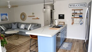 Una cocina moderna con una isla central, un área de comedor con un sofá gris, y una sala de estar con ventilador de techo.