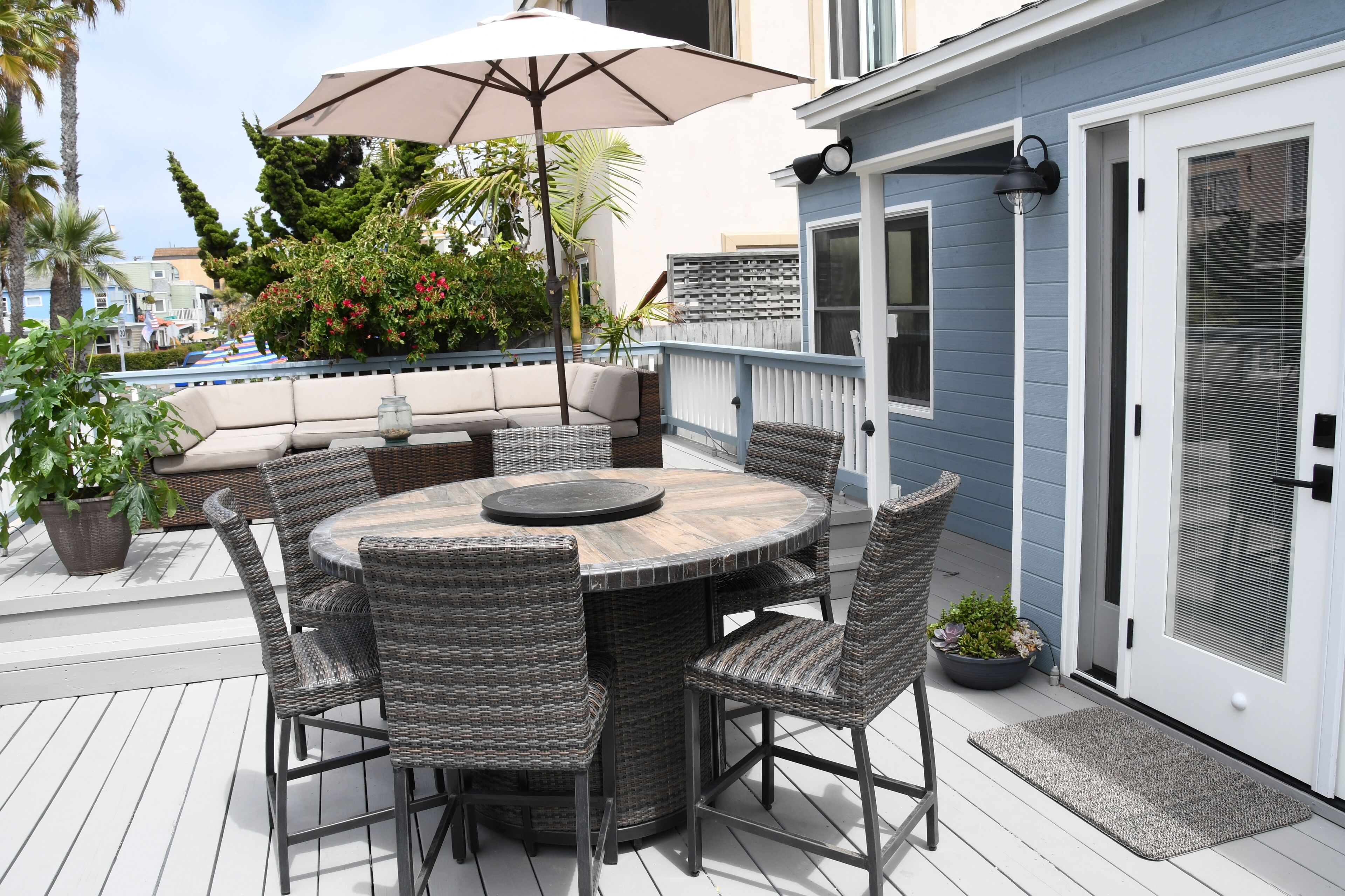 A deck with a round table and wicker chairs, surrounded by potted plants and a large umbrella.