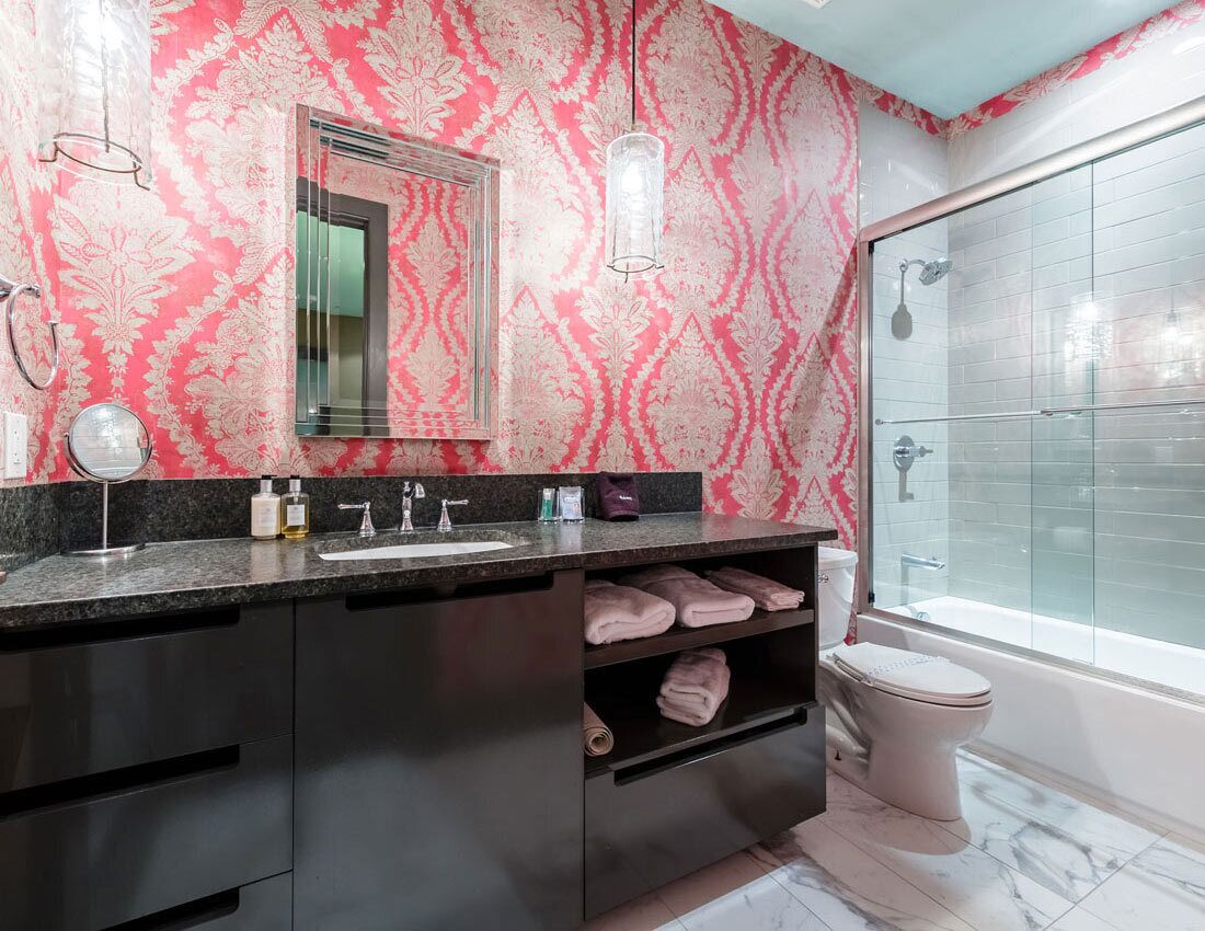 A bathroom with a pink patterned wall, a large mirror, a dark countertop, a toilet, and a glass-enclosed shower.