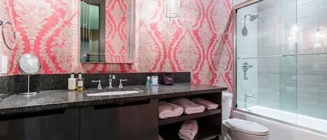 A bathroom with a pink patterned wall, a large mirror, a dark countertop, a toilet, and a glass-enclosed shower.