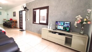 Een woonkamer met een bank, een televisie op een meubelstuk, een ventilator en een vaas met bloemen.