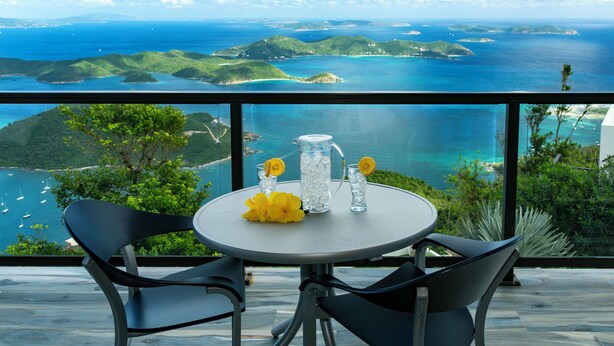 A table with chairs on a balcony overlooking a coastal landscape with clear skies and distant islands.