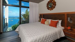 A bedroom with a large bed, a wooden headboard, a nightstand, and a view of the sea through a sliding door.