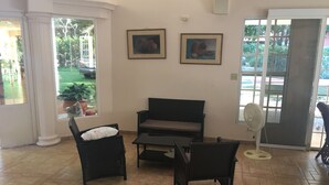 A room with a black sofa, two wicker chairs, a small table, and a standing fan. There are two framed pictures on the wall and a window with a view of greenery outside. - Panama Santa Clara Beach Residence (Cocle)