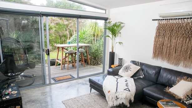 A modern living room with a black leather sofa, a glass sliding door leading to an outdoor area, and a large potted plant. -  BEACHWOOD STUDIO-a perfectly styled getaway for couples BEACH AT YOUR DOORSTEP (Smiths Beach)