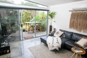 A modern living room with a black leather sofa, a glass sliding door leading to an outdoor area, and a large potted plant. - BEACHWOOD STUDIO YOUR BEACH ESCAPE STARTS AT YOUR DOORSTEP (Smiths Beach)