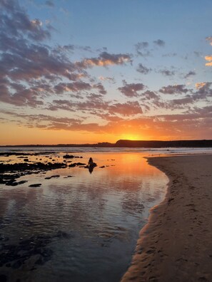 Une plage avec une personne assise sur les rochers, un ciel dégagé et un coucher de soleil.