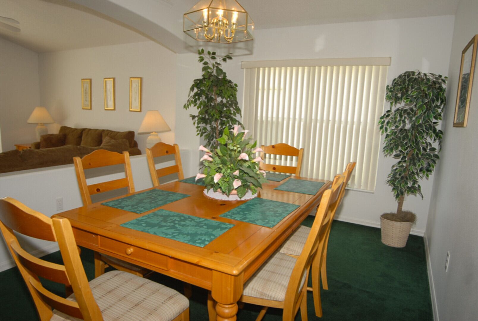 A dining area with a wooden table, chairs, and a central floral arrangement. There are potted plants on either side of the window, and a sofa in the background.