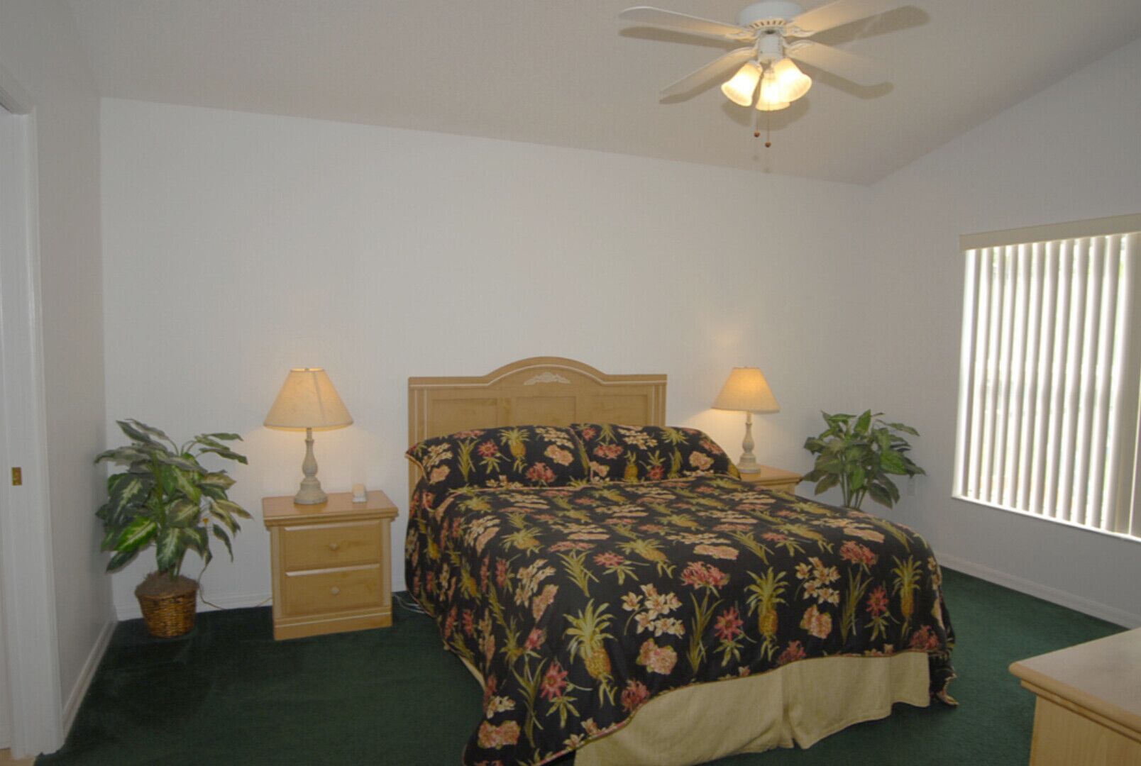 A bedroom with a bed, two bedside tables with lamps, a ceiling fan, a window with blinds, and two potted plants.