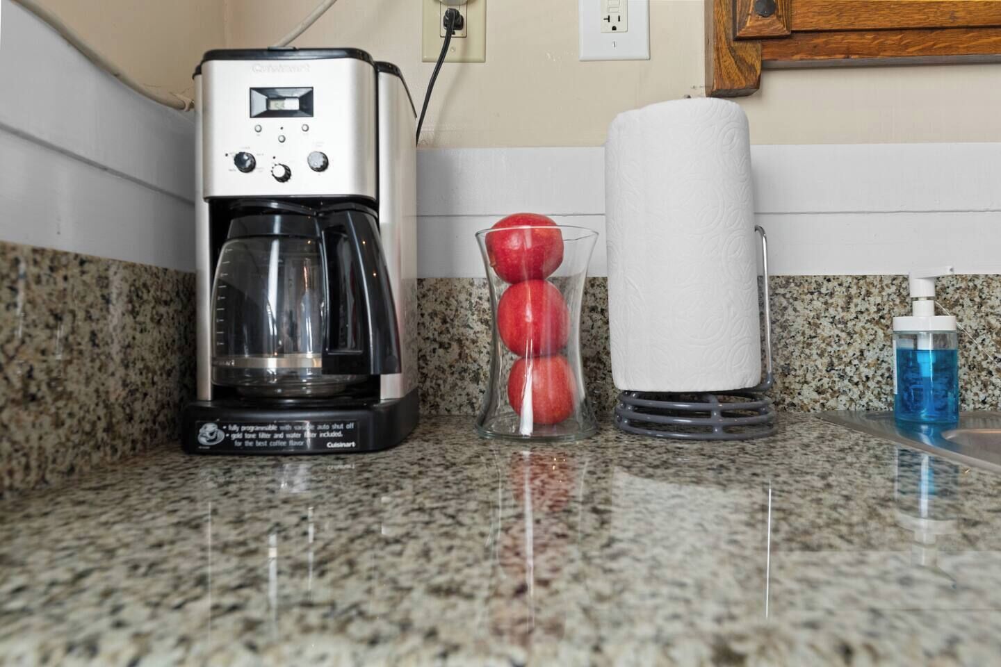 A kitchen counter with a coffee maker, a glass of red apples, a roll of paper towels, and a bottle of blue liquid soap.