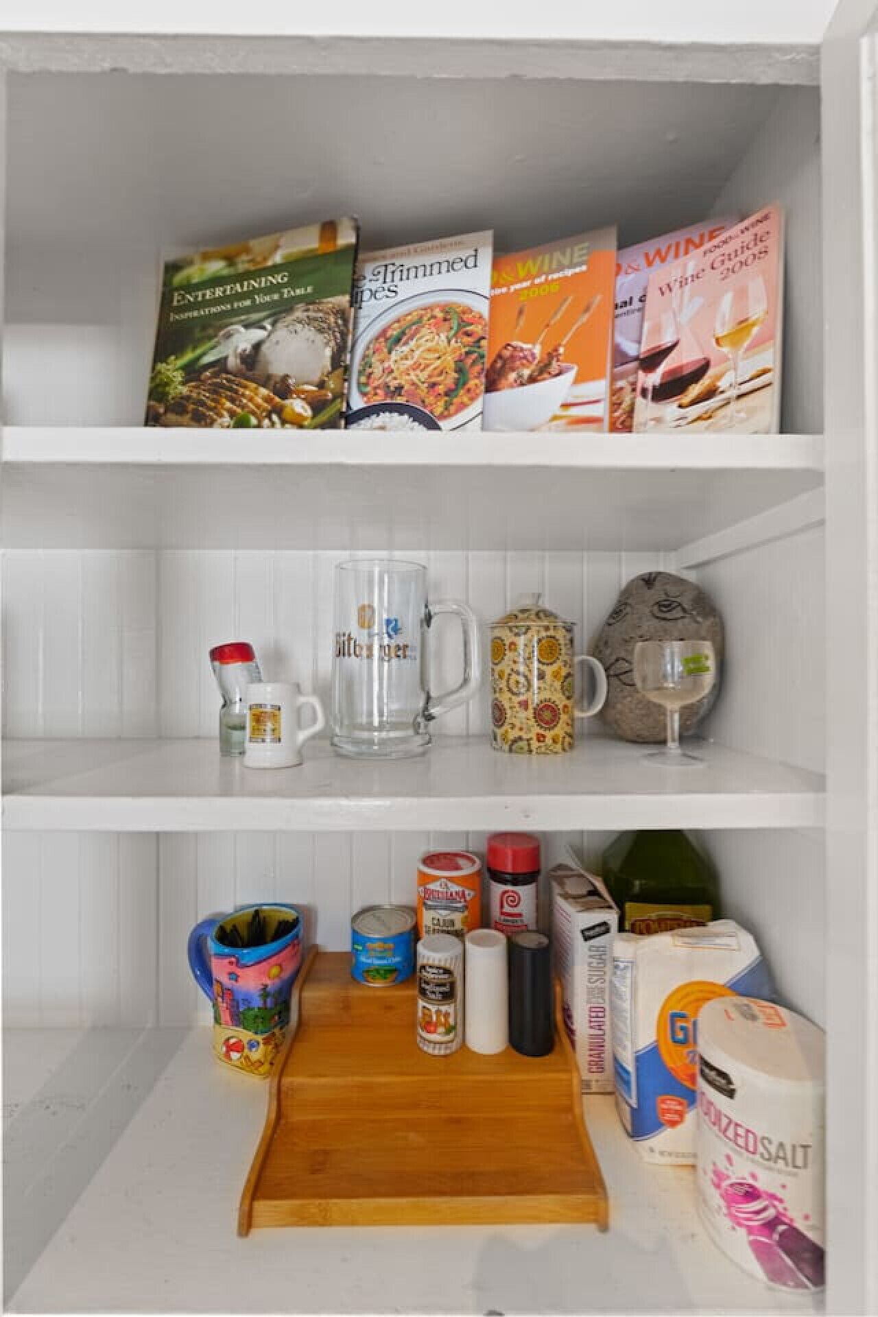 A kitchen shelf with cookbooks, a glass mug, a patterned mug, a wine glass, and various kitchen items like a rolling pin, a can, and a bottle of sugar.