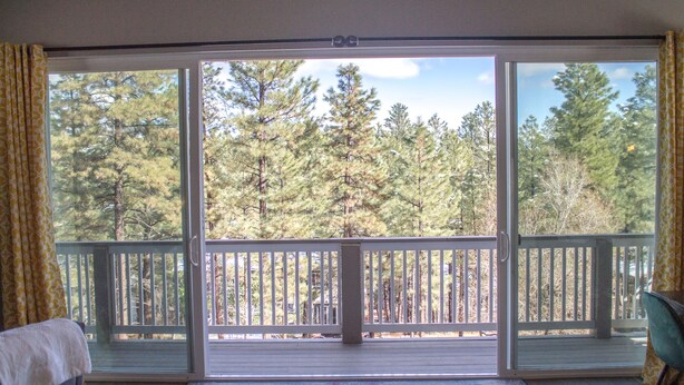 A room with a sliding glass door leading to a balcony with a wooden railing, a patterned rug on the floor, and a chair on the right side.