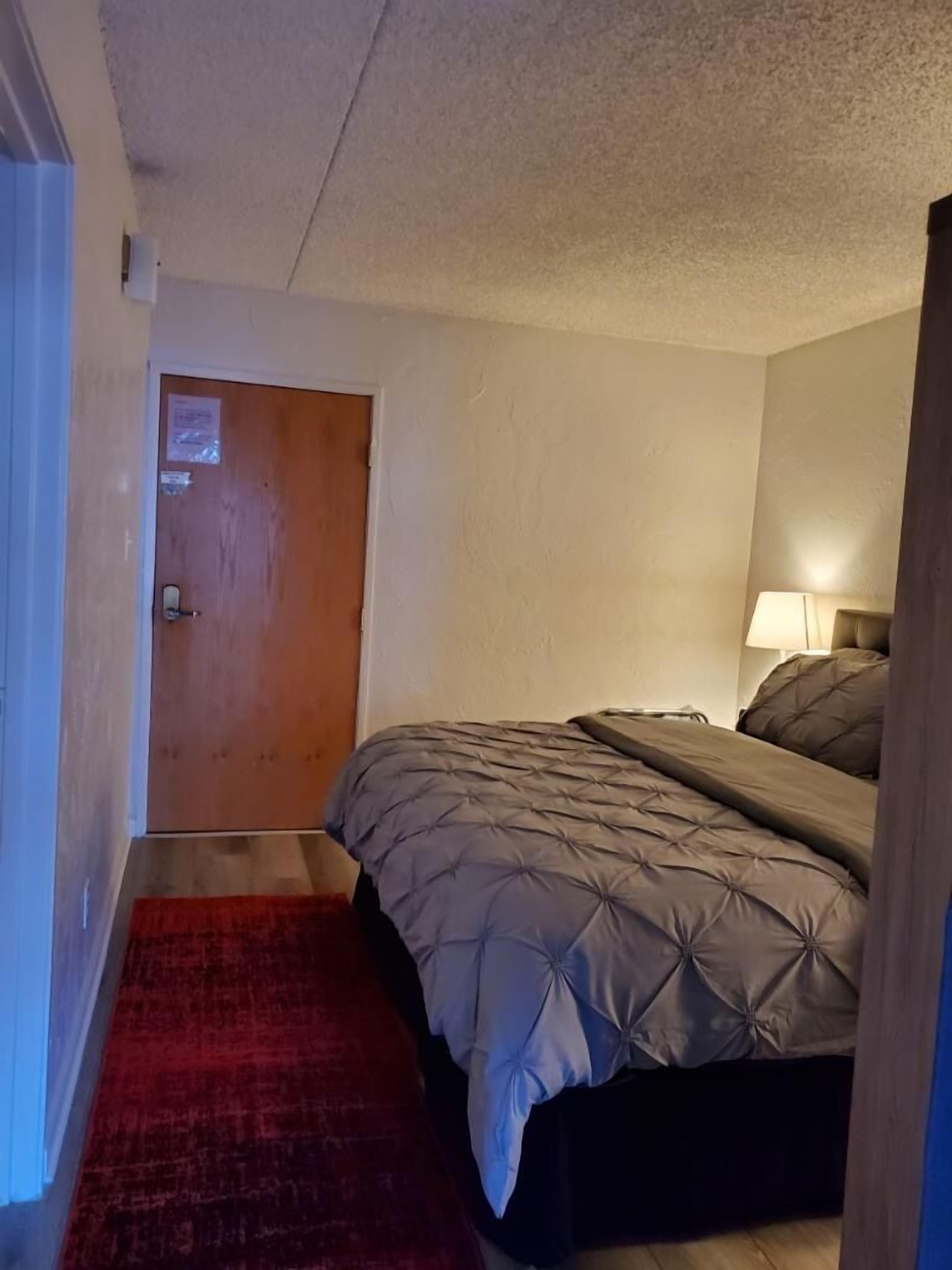 Un dormitorio con una cama, un tapete rojo, una puerta de madera y una mesita de noche con una lámpara.